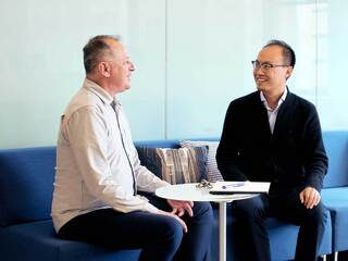 A smiling man with glasses sits at a table with a laptop, engaged in conversation with a man across from him.