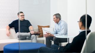 Three professionals engage in a discussion, sharing insights on laptops while seated comfortably in a modern, well-lit meeting space.