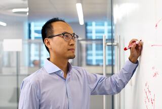 A focused man writes on a transparent board, deep in thought, surrounded by a bright, modern workspace with large windows.